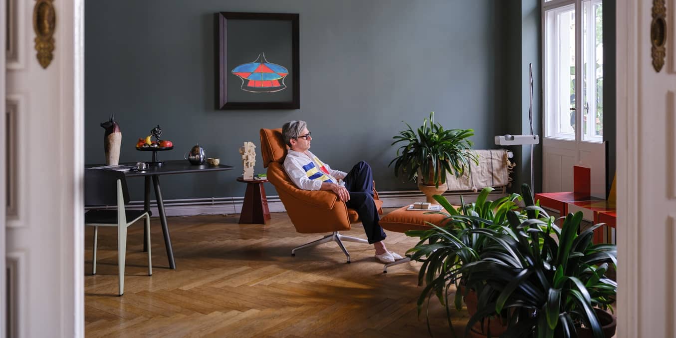 Orangefarbener Lounge-Sessel mit Ottoman auf Fischgr&auml;tparkett vor blaugrauer Wand, umgeben von Zimmerpflanzen